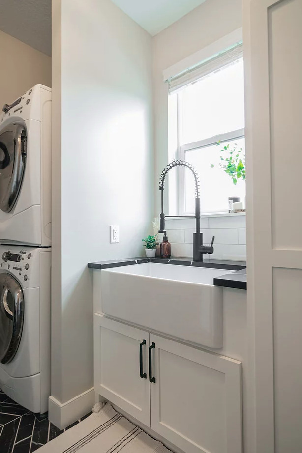 This is an example of a mid-sized transitional galley utility room in Minneapolis with a farmhouse sink, shaker cabinets, white cabinets, quartz benchtops, white splashback, ceramic splashback, grey walls, ceramic floors, a stacked washer and dryer, black floor and black benchtop.