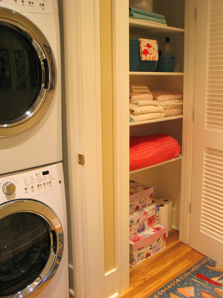 Inspiration for a mid-sized traditional laundry cupboard in Portland with louvered cabinets, white cabinets, yellow walls, medium hardwood floors, a stacked washer and dryer and brown floor.