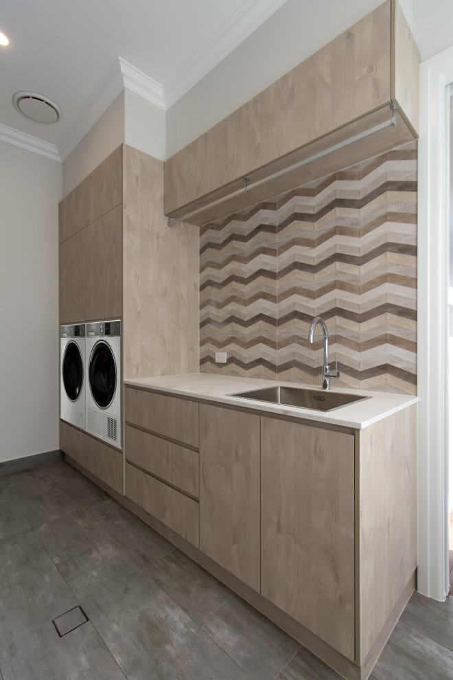 Inspiration for a small contemporary single-wall dedicated laundry room in Sydney with a drop-in sink, flat-panel cabinets, light wood cabinets, quartz benchtops, multi-coloured splashback, porcelain splashback, beige walls, porcelain floors, a side-by-side washer and dryer, grey floor and white benchtop.