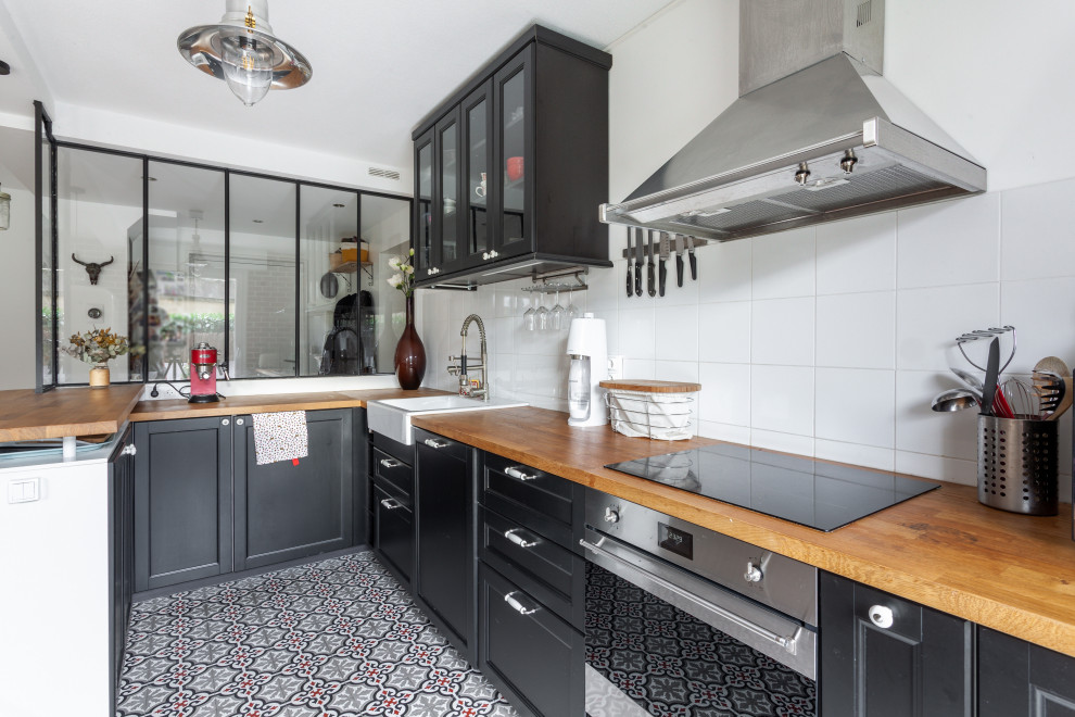 This is an example of a mid-sized transitional u-shaped kitchen in Lyon with a farmhouse sink, recessed-panel cabinets, black cabinets, wood benchtops, white splashback, porcelain splashback, stainless steel appliances, ceramic floors, a peninsula, multi-coloured floor and beige benchtop.