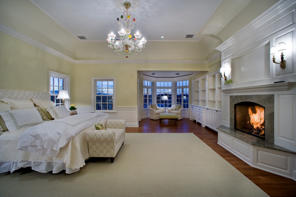 Photo of a large traditional master bedroom in New York with yellow walls, medium hardwood floors, a standard fireplace, a stone fireplace surround and brown floor.