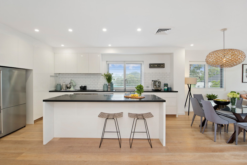 Design ideas for a modern single-wall eat-in kitchen in Sydney with white splashback, stainless steel appliances, light hardwood floors, with island, a drop-in sink, flat-panel cabinets, white cabinets, granite benchtops, subway tile splashback and black benchtop.