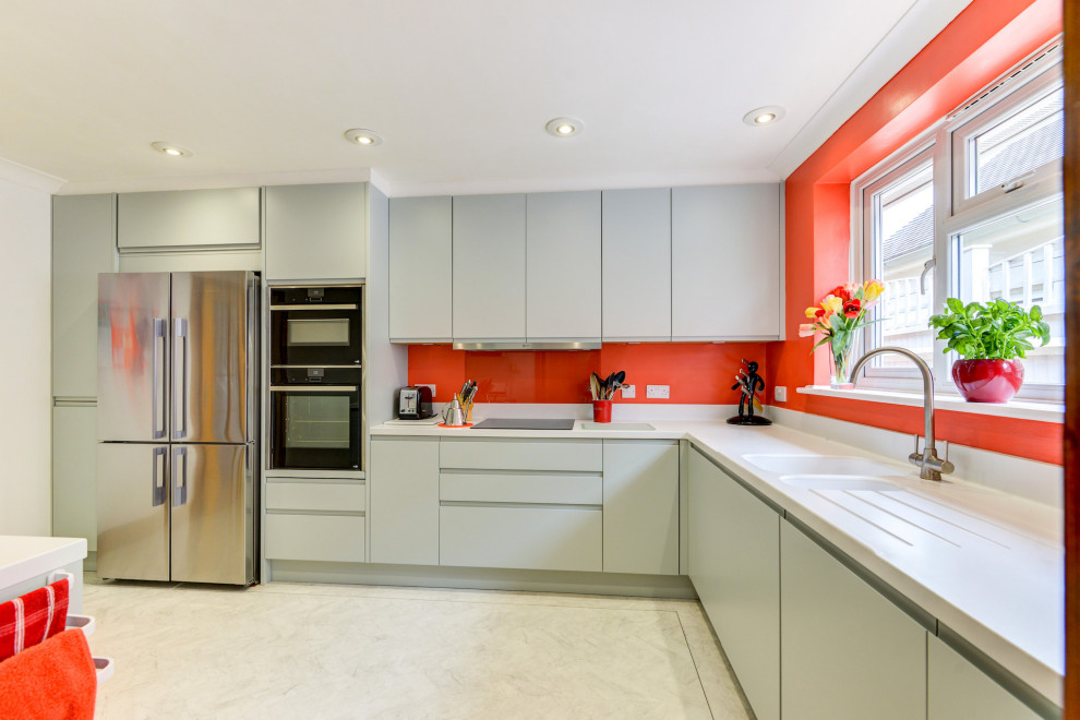 Mid-sized beach style l-shaped separate kitchen in Sussex with a drop-in sink, flat-panel cabinets, green cabinets, solid surface benchtops, orange splashback, glass sheet splashback, black appliances, linoleum floors, a peninsula, white floor and white benchtop.