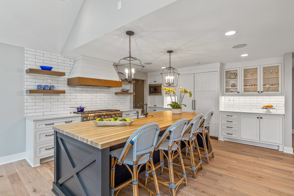 Design ideas for a country l-shaped kitchen in Cleveland with a farmhouse sink, beaded inset cabinets, white cabinets, white splashback, subway tile splashback, stainless steel appliances, medium hardwood floors, with island, brown floor and white benchtop.