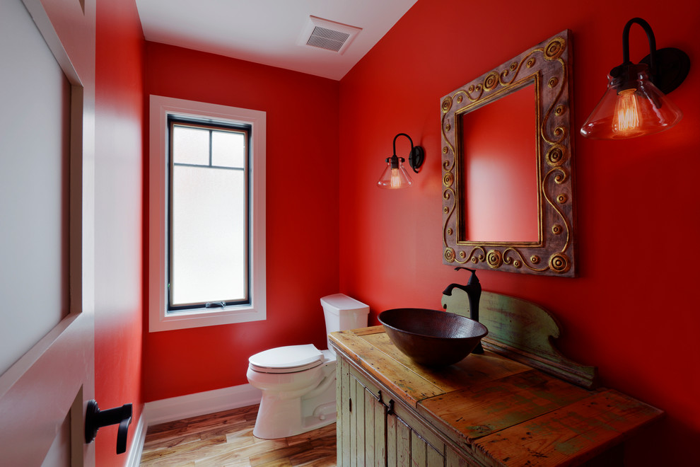 Photo of a mid-sized arts and crafts powder room in Ottawa with red walls, louvered cabinets, distressed cabinets, a two-piece toilet, light hardwood floors, a vessel sink, wood benchtops and brown benchtops.