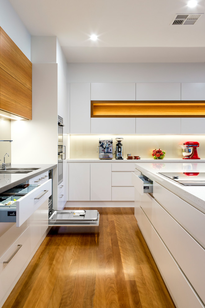 Photo of a large contemporary l-shaped separate kitchen in Adelaide with an integrated sink, flat-panel cabinets, white cabinets, quartz benchtops, metallic splashback, glass sheet splashback, black appliances, medium hardwood floors, with island and white benchtop.