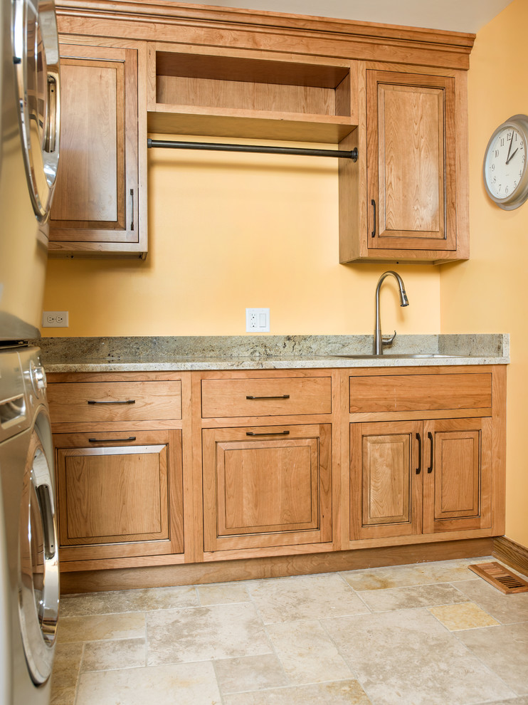 Photo of a large country l-shaped utility room in Chicago with an undermount sink, raised-panel cabinets, medium wood cabinets, granite benchtops, yellow walls, limestone floors and a stacked washer and dryer.