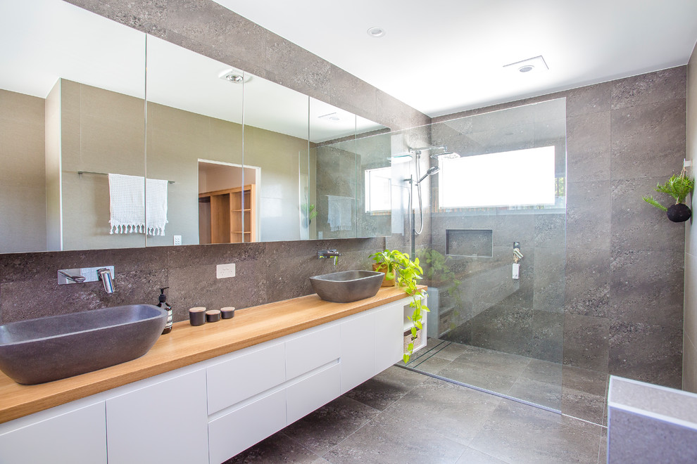 This is an example of a large contemporary master wet room bathroom in Sunshine Coast with flat-panel cabinets, white cabinets, stone tile, grey walls, ceramic floors, a vessel sink, wood benchtops, grey floor, gray tile, an open shower and brown benchtops.