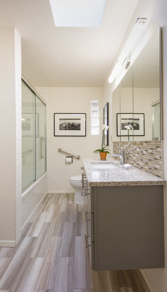 Photo of a small transitional 3/4 bathroom in San Luis Obispo with flat-panel cabinets, grey cabinets, an alcove tub, a shower/bathtub combo, beige tile, porcelain tile, an undermount sink, engineered quartz benchtops, a two-piece toilet, beige walls, light hardwood floors, beige floor and a sliding shower screen.