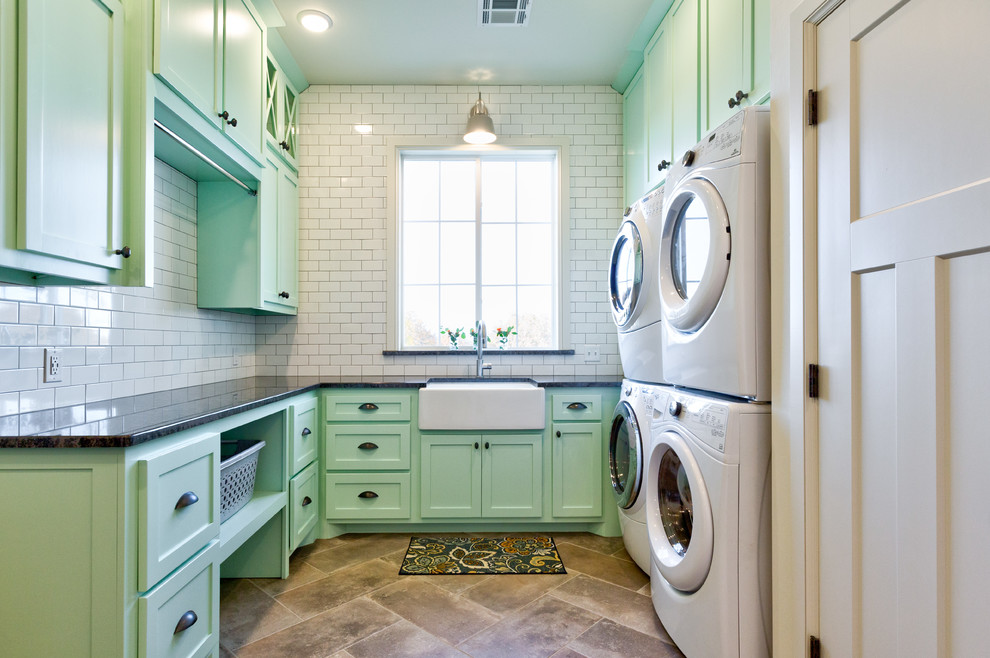 This is an example of a country u-shaped dedicated laundry room in Oklahoma City with a farmhouse sink, shaker cabinets, green cabinets, a stacked washer and dryer, brown floor and black benchtop.