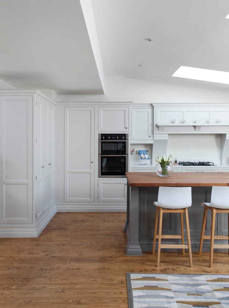 Large transitional l-shaped open plan kitchen in Dublin with a farmhouse sink, beaded inset cabinets, grey cabinets, quartzite benchtops, grey splashback, stone slab splashback, stainless steel appliances, medium hardwood floors, with island and brown floor.