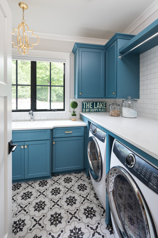 This is an example of a mid-sized beach style l-shaped dedicated laundry room in Minneapolis with an undermount sink, blue cabinets, solid surface benchtops, ceramic floors, multi-coloured floor, white benchtop, shaker cabinets, grey walls and a side-by-side washer and dryer.
