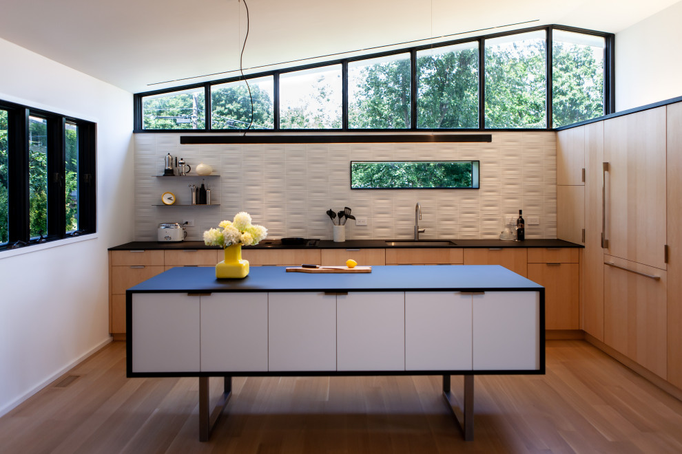 Inspiration for a mid-sized midcentury l-shaped open plan kitchen in New York with an undermount sink, flat-panel cabinets, light wood cabinets, laminate benchtops, white splashback, ceramic splashback, black appliances, light hardwood floors, with island and black benchtop.