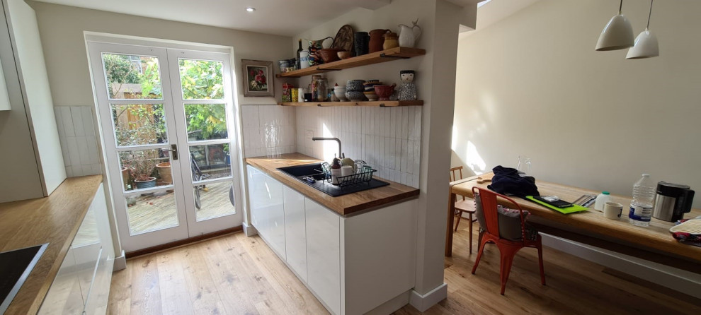 Inspiration for a small modern galley separate kitchen in Oxfordshire with an integrated sink, flat-panel cabinets, white cabinets, wood benchtops, white splashback, porcelain splashback, black appliances, medium hardwood floors, brown floor and brown benchtop.