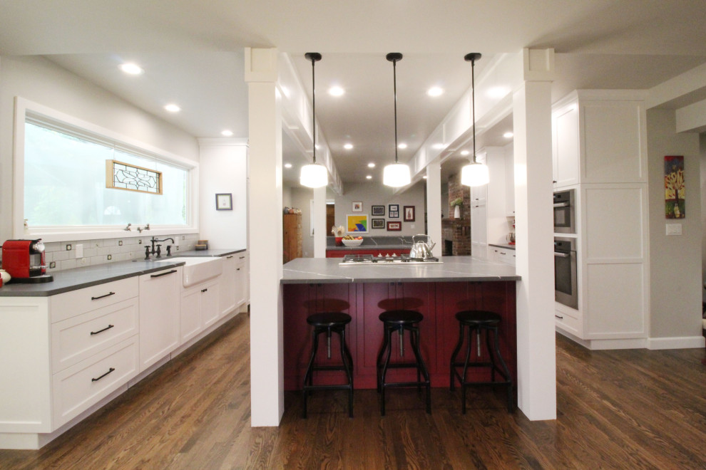 This is an example of a mid-sized country galley open plan kitchen in Seattle with a farmhouse sink, shaker cabinets, red cabinets, quartz benchtops, grey splashback, cement tile splashback, panelled appliances, medium hardwood floors, multiple islands, brown floor, grey benchtop and exposed beam.