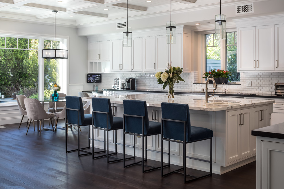 Photo of a beach style eat-in kitchen in Los Angeles with recessed-panel cabinets, white cabinets, white splashback, subway tile splashback, dark hardwood floors, with island, brown floor and black benchtop.