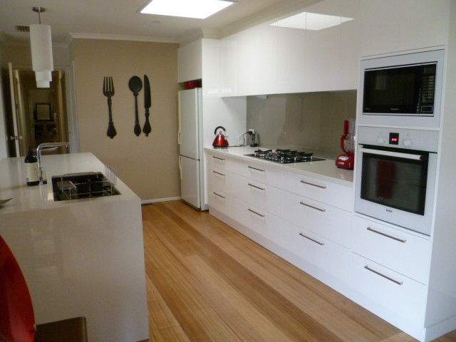 Photo of a mid-sized modern galley kitchen pantry in Melbourne with an undermount sink, white cabinets, granite benchtops, metallic splashback, glass sheet splashback, white appliances, light hardwood floors and with island.