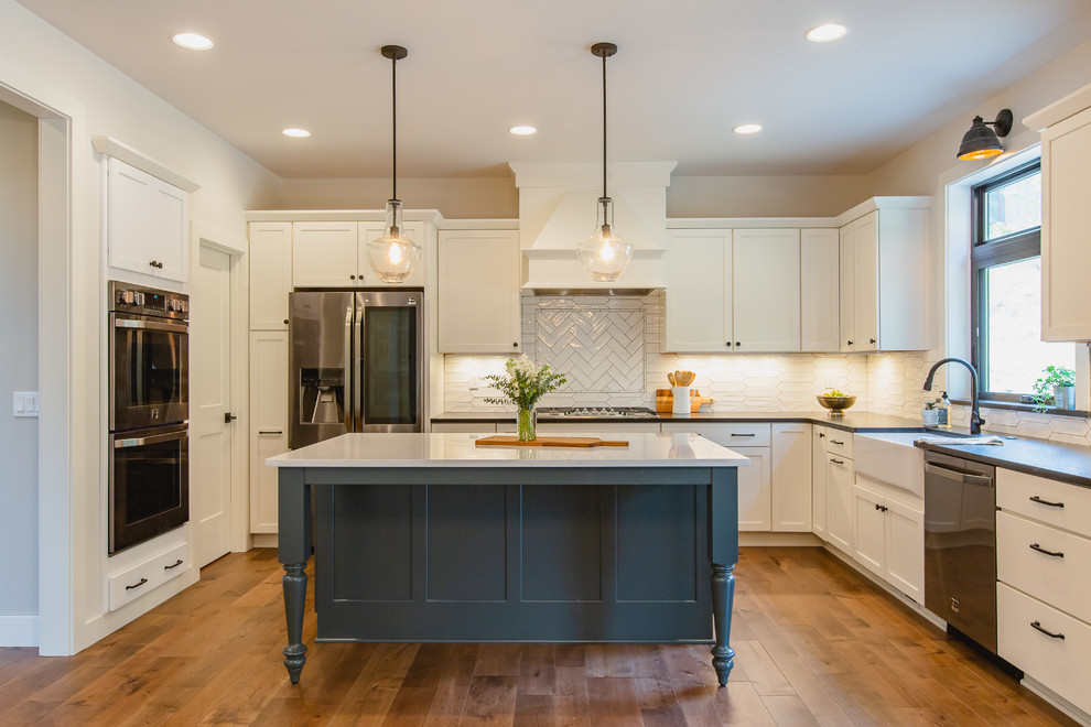 Design ideas for a mid-sized country u-shaped open plan kitchen in Seattle with a farmhouse sink, shaker cabinets, white cabinets, granite benchtops, white splashback, ceramic splashback, stainless steel appliances, medium hardwood floors, with island, black benchtop and brown floor.