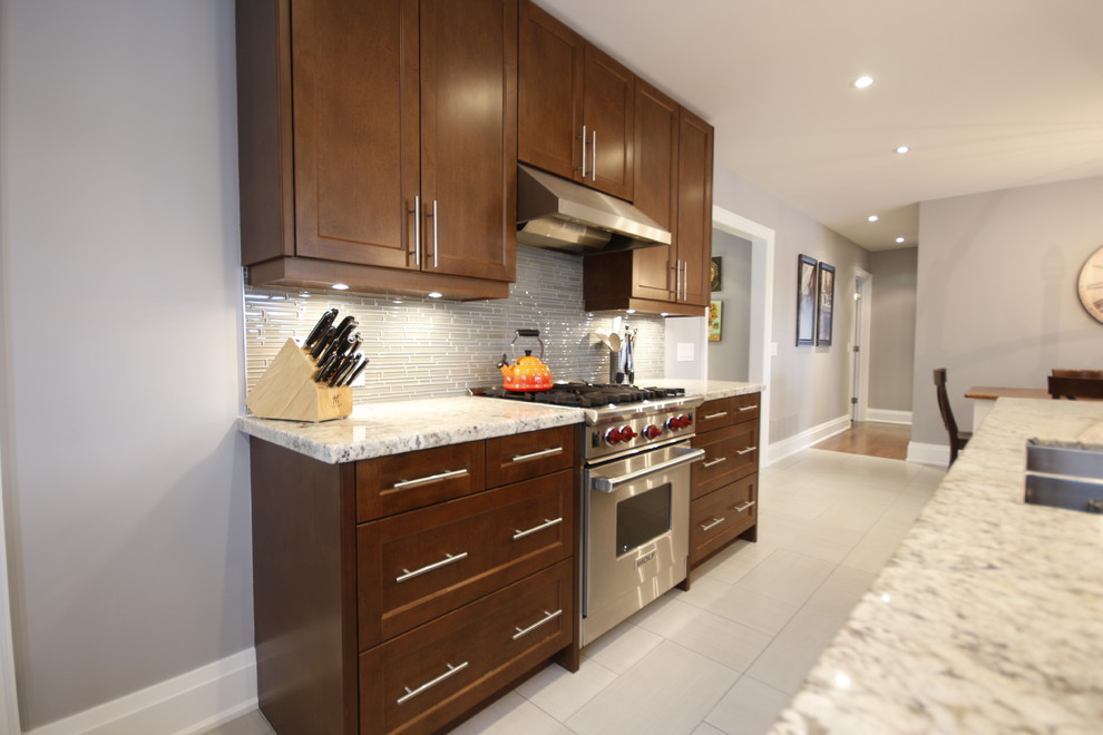 Photo of a mid-sized transitional l-shaped eat-in kitchen in Toronto with an undermount sink, recessed-panel cabinets, brown cabinets, granite benchtops, beige splashback, matchstick tile splashback, stainless steel appliances, porcelain floors, with island and beige floor.