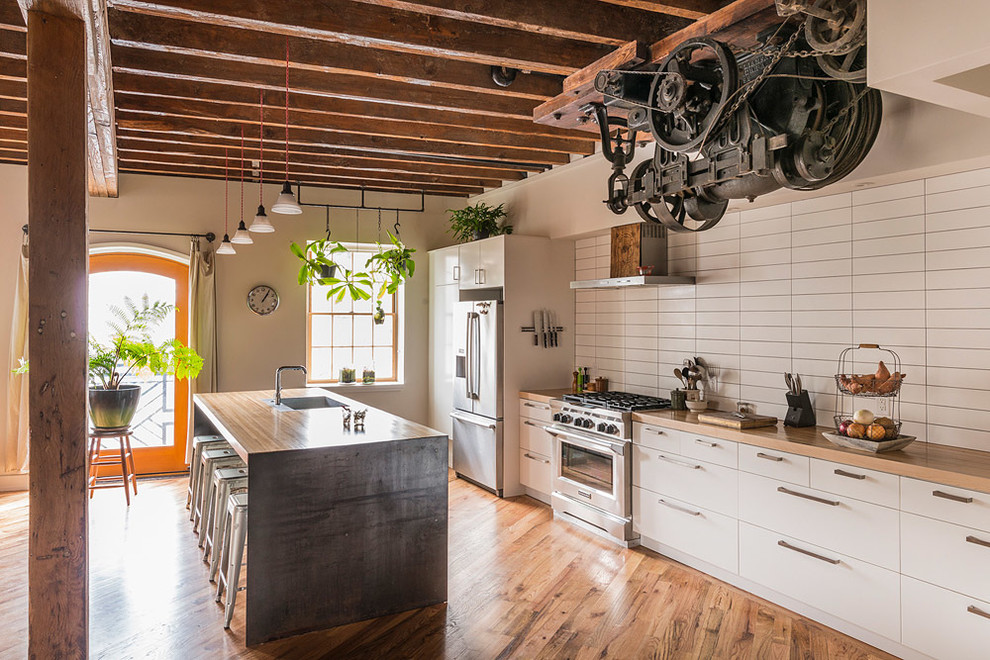 This is an example of a large industrial kitchen in Philadelphia with flat-panel cabinets, white cabinets, wood benchtops, white splashback, stainless steel appliances, with island, ceramic splashback, a single-bowl sink, medium hardwood floors and brown floor.