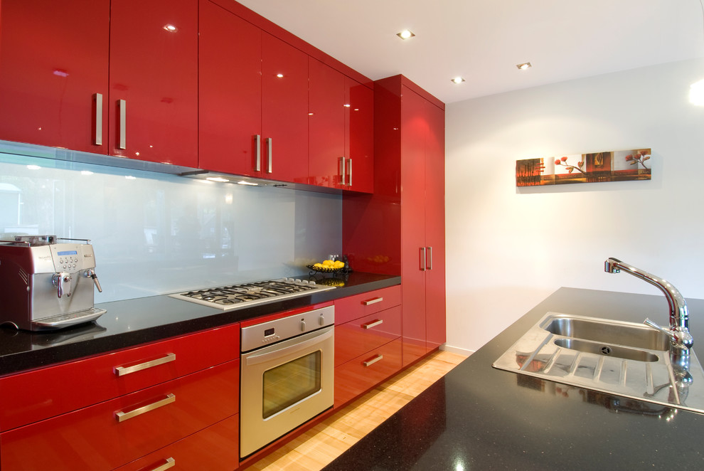 Photo of a mid-sized contemporary galley open plan kitchen in Auckland with a double-bowl sink, flat-panel cabinets, red cabinets, quartz benchtops, blue splashback, glass sheet splashback, stainless steel appliances, medium hardwood floors, with island and yellow floor.