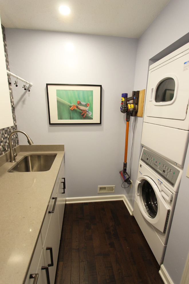 This is an example of a small transitional galley dedicated laundry room in Cleveland with an undermount sink, white cabinets, quartz benchtops, grey walls, medium hardwood floors, a stacked washer and dryer, brown floor and beige benchtop.
