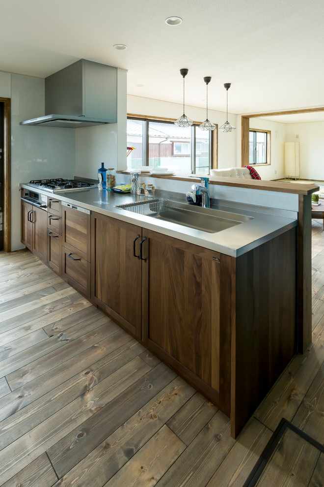 This is an example of a country single-wall open plan kitchen in Other with an integrated sink, recessed-panel cabinets, distressed cabinets, stainless steel benchtops, medium hardwood floors, a peninsula and brown floor.