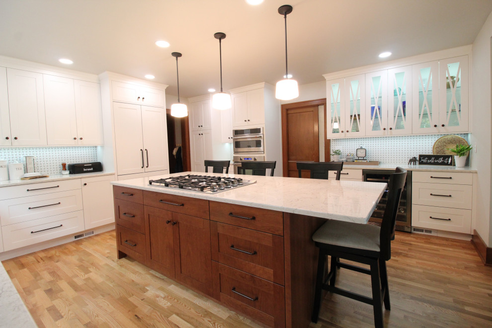 Photo of a mid-sized traditional l-shaped eat-in kitchen in Seattle with a farmhouse sink, recessed-panel cabinets, white cabinets, quartz benchtops, white splashback, ceramic splashback, stainless steel appliances, light hardwood floors, with island, brown floor and white benchtop.