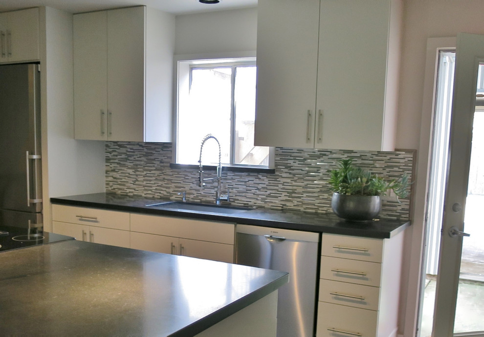 Photo of a small industrial single-wall open plan kitchen in Houston with a farmhouse sink, flat-panel cabinets, white cabinets, granite benchtops, grey splashback, porcelain splashback, stainless steel appliances, concrete floors, with island, grey floor, black benchtop and exposed beam.