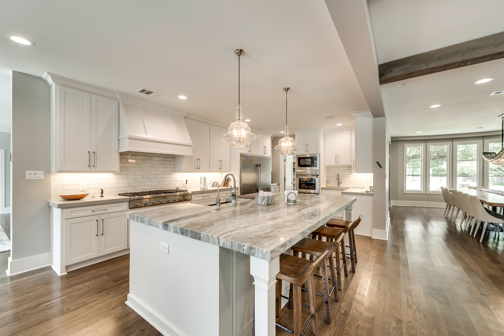 Huge transitional l-shaped medium tone wood floor and brown floor open concept kitchen photo in Dallas with a farmhouse sink, shaker cabinets, white cabinets, quartz countertops, gray backsplash, subway tile backsplash, stainless steel appliances, an island and multicolored countertops