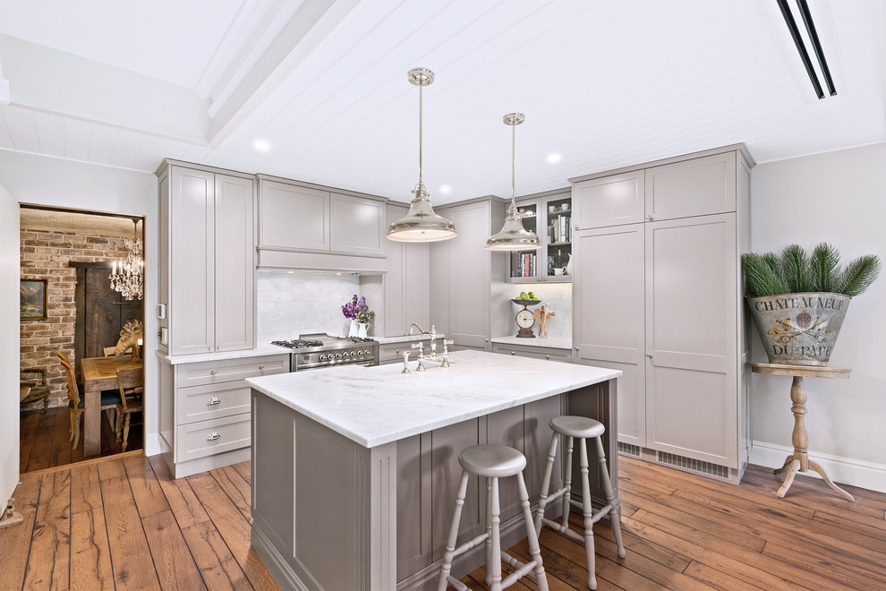 Transitional l-shaped kitchen in Sydney with a farmhouse sink, recessed-panel cabinets, grey cabinets, white splashback, stainless steel appliances, medium hardwood floors, with island, brown floor and white benchtop.