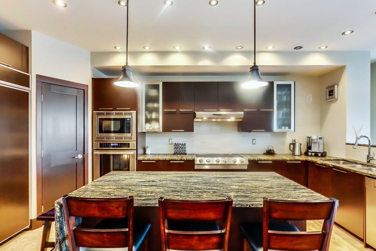 Large transitional l-shaped travertine floor open concept kitchen photo in Calgary with a double-bowl sink, flat-panel cabinets, dark wood cabinets, granite countertops, beige backsplash, stainless steel appliances and an island