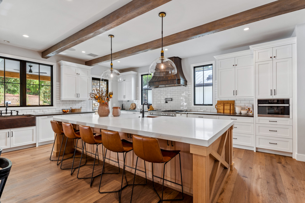 Photo of an expansive country l-shaped open plan kitchen in Los Angeles with a farmhouse sink, shaker cabinets, white cabinets, quartz benchtops, white splashback, ceramic splashback, stainless steel appliances, light hardwood floors, with island, beige floor, multi-coloured benchtop and exposed beam.