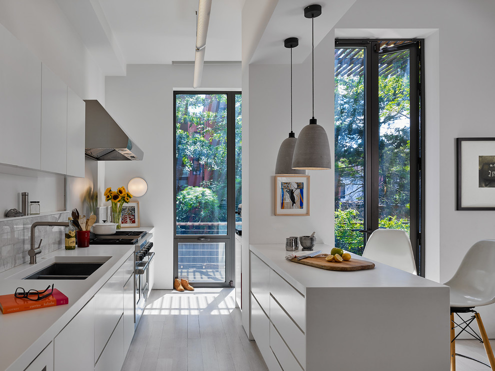 Photo of a mid-sized contemporary galley kitchen in New York with an undermount sink, flat-panel cabinets, white cabinets, stainless steel appliances, with island, white benchtop, grey splashback, painted wood floors and white floor.