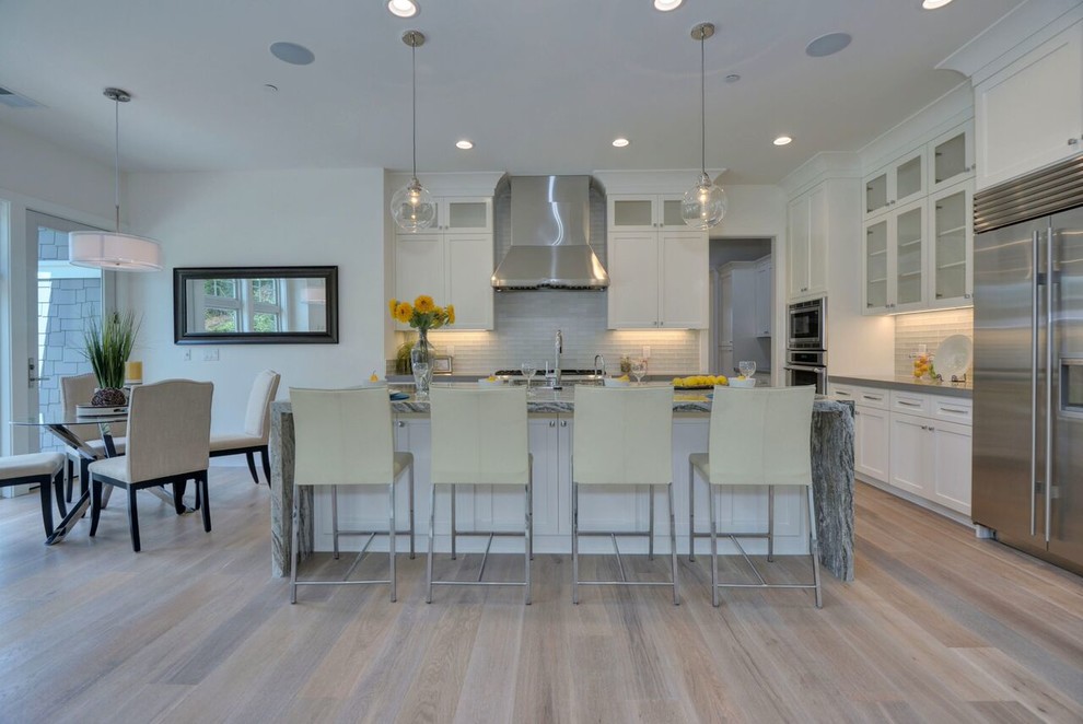 Design ideas for a large transitional l-shaped open plan kitchen in San Francisco with a farmhouse sink, shaker cabinets, white cabinets, quartz benchtops, glass tile splashback, stainless steel appliances, light hardwood floors, with island, brown floor and grey benchtop.