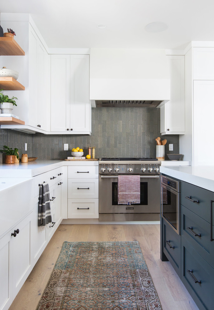 Photo of a mid-sized transitional l-shaped kitchen in San Diego with a farmhouse sink, shaker cabinets, white cabinets, solid surface benchtops, grey splashback, subway tile splashback, panelled appliances, light hardwood floors, with island and beige floor.