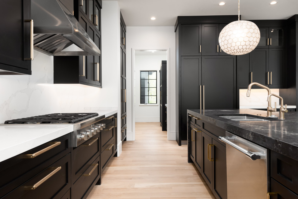 Example of a mid-sized transitional u-shaped light wood floor kitchen design with an undermount sink, shaker cabinets, black cabinets, quartz countertops, white backsplash, quartz backsplash, stainless steel appliances, an island and black countertops