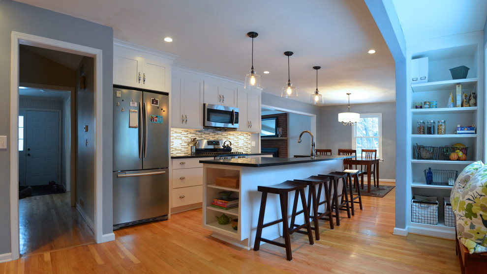 Example of a mid-sized transitional single-wall medium tone wood floor eat-in kitchen design in New York with a farmhouse sink, shaker cabinets, white cabinets, granite countertops, gray backsplash, glass tile backsplash, stainless steel appliances and an island
