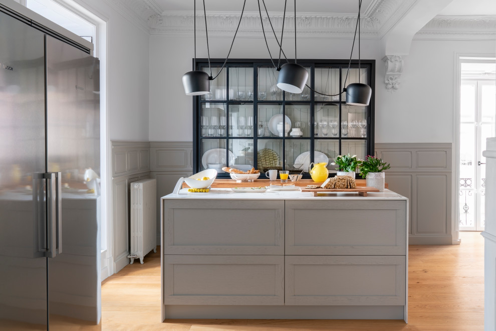 Aménagement d'une cuisine américaine linéaire et grise et blanche de taille moyenne avec un placard avec porte à panneau surélevé, des portes de placard grises, un plan de travail en quartz modifié, une crédence blanche, une crédence en quartz modifié, un électroménager en acier inoxydable, un sol en bois brun, îlot et un plan de travail blanc.