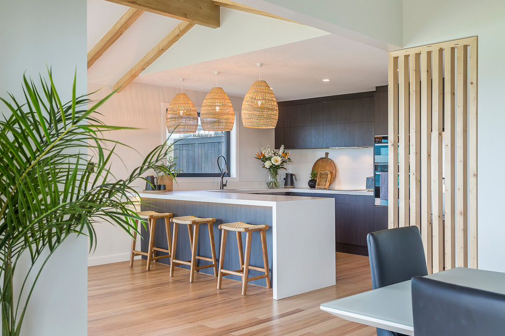 This is an example of a mid-sized beach style u-shaped open plan kitchen in Auckland with an undermount sink, flat-panel cabinets, dark wood cabinets, quartz benchtops, white splashback, porcelain splashback, stainless steel appliances, light hardwood floors, a peninsula and white benchtop.