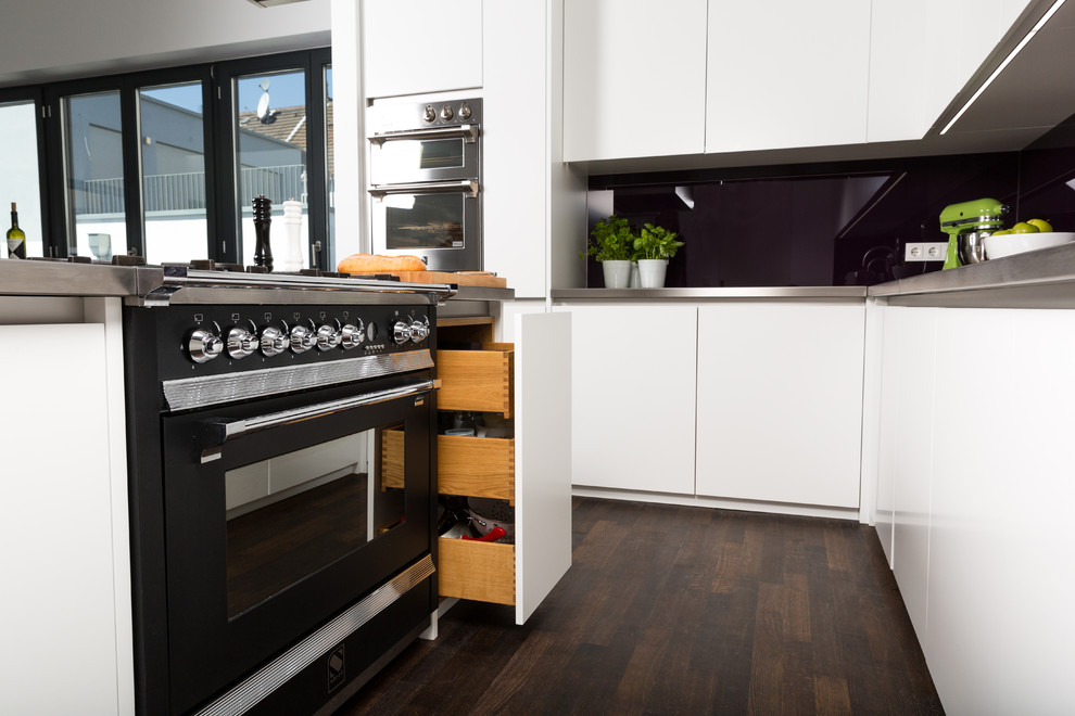 Design ideas for a large industrial open plan kitchen in Cologne with a double-bowl sink, flat-panel cabinets, white cabinets, stainless steel benchtops, pink splashback, glass sheet splashback, coloured appliances, dark hardwood floors and with island.