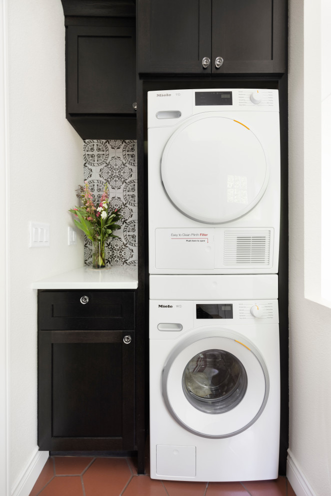 This is an example of a small transitional utility room in Portland with shaker cabinets, black cabinets, quartz benchtops, multi-coloured splashback, terra-cotta splashback, white walls, ceramic floors, a stacked washer and dryer, red floor and white benchtop.
