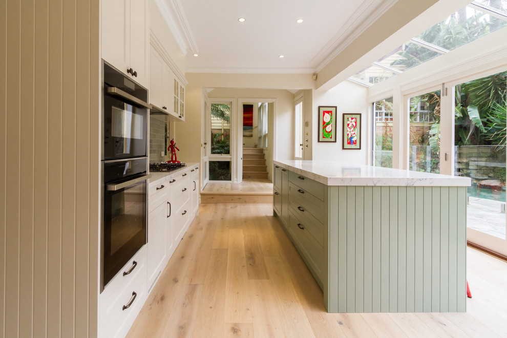 Design ideas for a mid-sized traditional galley eat-in kitchen in Melbourne with an undermount sink, shaker cabinets, white cabinets, quartz benchtops, green splashback, subway tile splashback, black appliances, light hardwood floors, with island, beige floor and white benchtop.