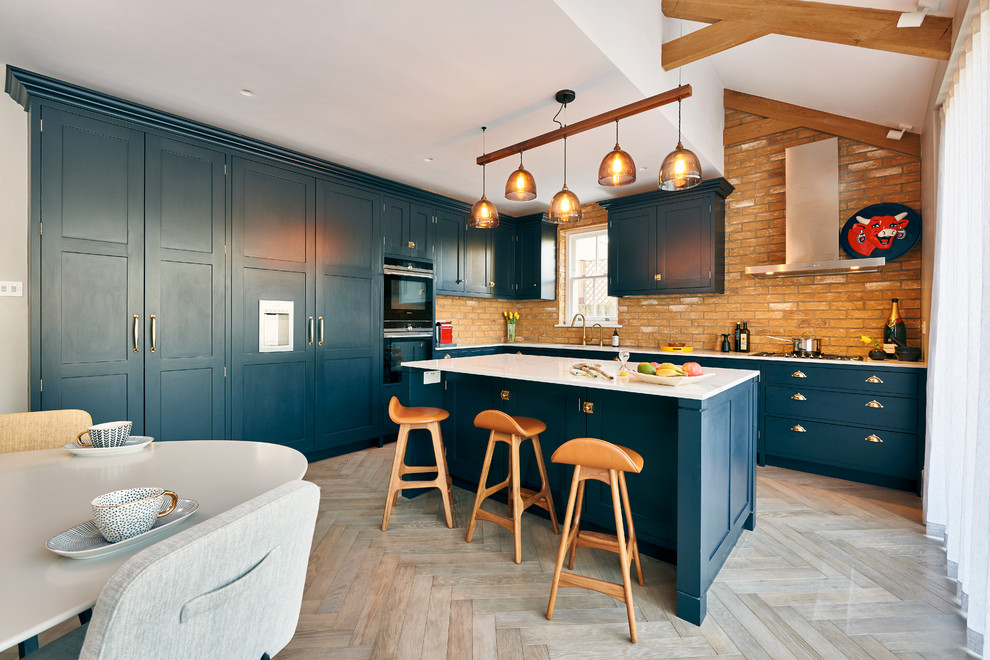 Design ideas for a large eclectic l-shaped open plan kitchen in London with an integrated sink, shaker cabinets, blue cabinets, quartz benchtops, brick splashback, panelled appliances, medium hardwood floors, with island and white benchtop.