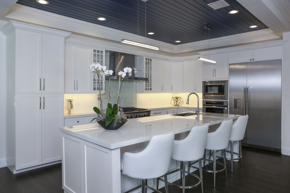 This is an example of a mid-sized beach style kitchen in Miami with a drop-in sink, shaker cabinets, white cabinets, quartz benchtops, white splashback, engineered quartz splashback, stainless steel appliances, with island and white benchtop.