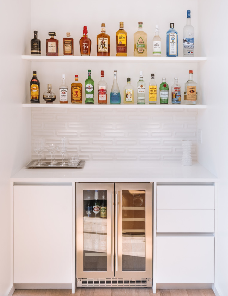 This is an example of a modern single-wall home bar in Portland with flat-panel cabinets, white cabinets, white splashback, light hardwood floors, brown floor and white benchtop.