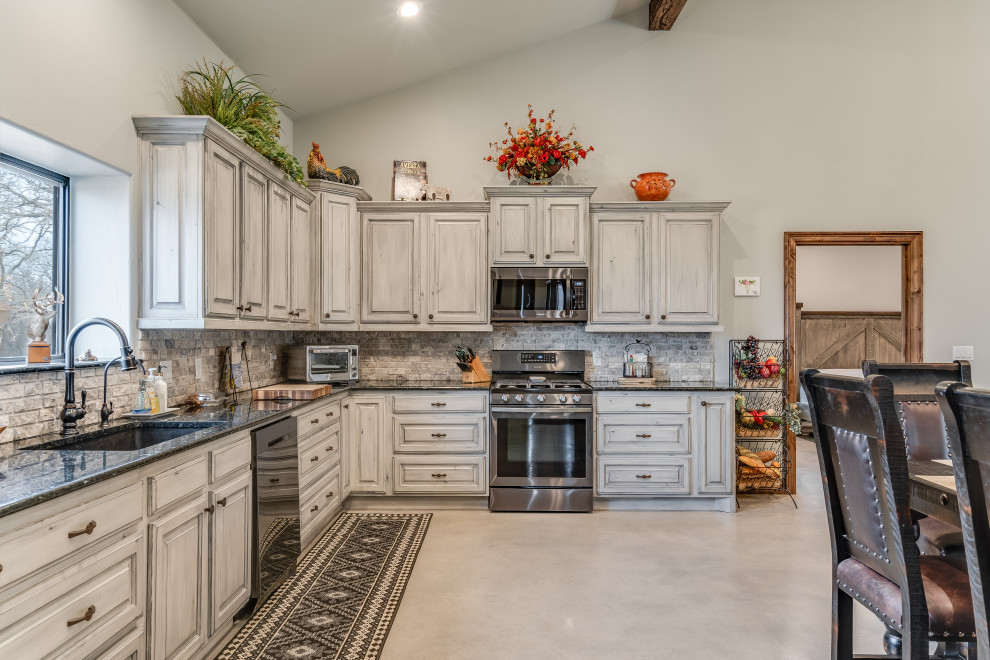 This is an example of a mid-sized country l-shaped eat-in kitchen in Austin with an undermount sink, raised-panel cabinets, grey cabinets, granite benchtops, grey splashback, stone tile splashback, stainless steel appliances, concrete floors, grey floor, black benchtop and vaulted.