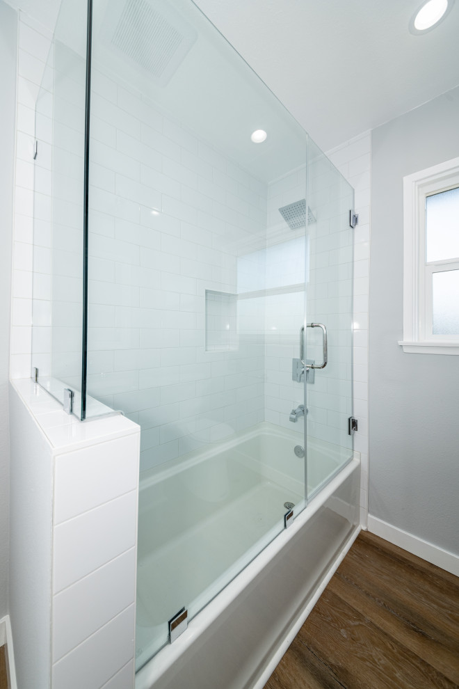 This is an example of a medium sized traditional grey and white shower room bathroom in Los Angeles with shaker cabinets, grey cabinets, a submerged bath, a shower/bath combination, a one-piece toilet, white tiles, metro tiles, white walls, laminate floors, a submerged sink, marble worktops, brown floors, a hinged door, white worktops, a wall niche, a single sink and a freestanding vanity unit.