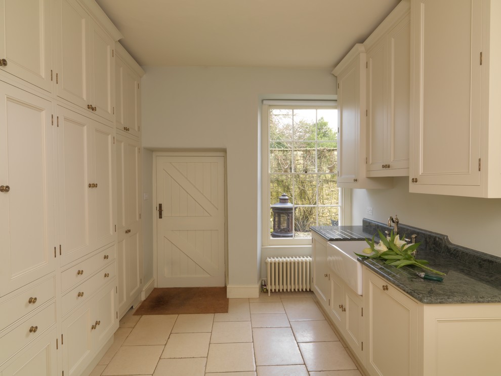 Photo of a mid-sized traditional galley utility room in Other with a farmhouse sink, recessed-panel cabinets, white cabinets, granite benchtops, white walls, limestone floors and a concealed washer and dryer.