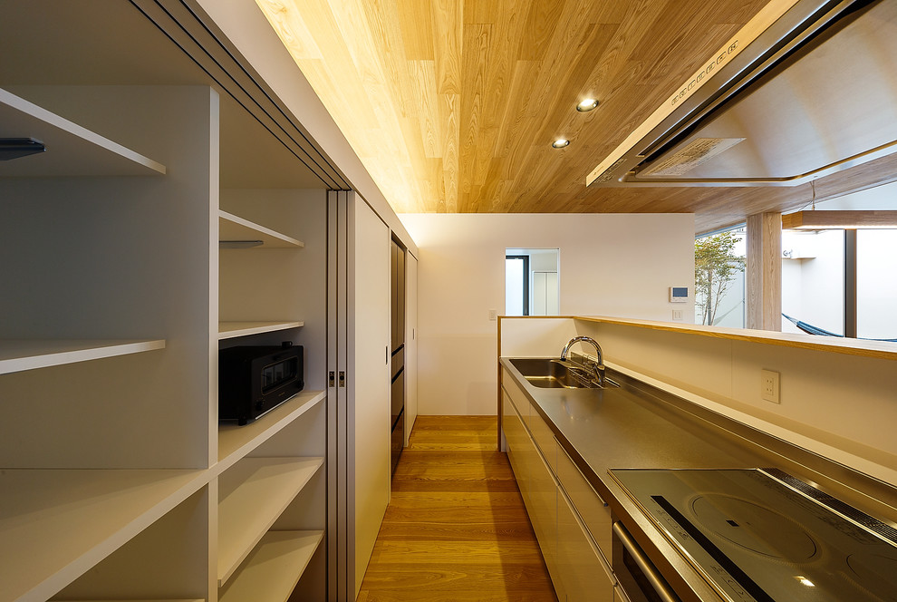Mid-sized modern single-wall open plan kitchen in Other with flat-panel cabinets, white cabinets, stainless steel benchtops, white splashback, plywood floors, a peninsula and brown floor.